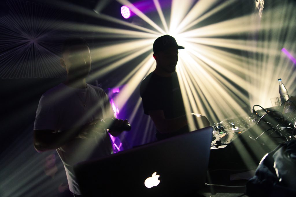 Ein DJ spielt an einem Deck mit Apple Laptop und wird mit einen Scheinwerfer von hinten bestrahlt bei einem event Veranstaltung Party